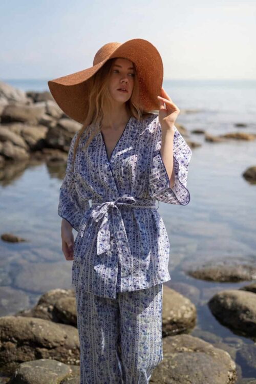 Woman in patterned beachwear with wide-brim hat by rocky shoreline on cloudy day.