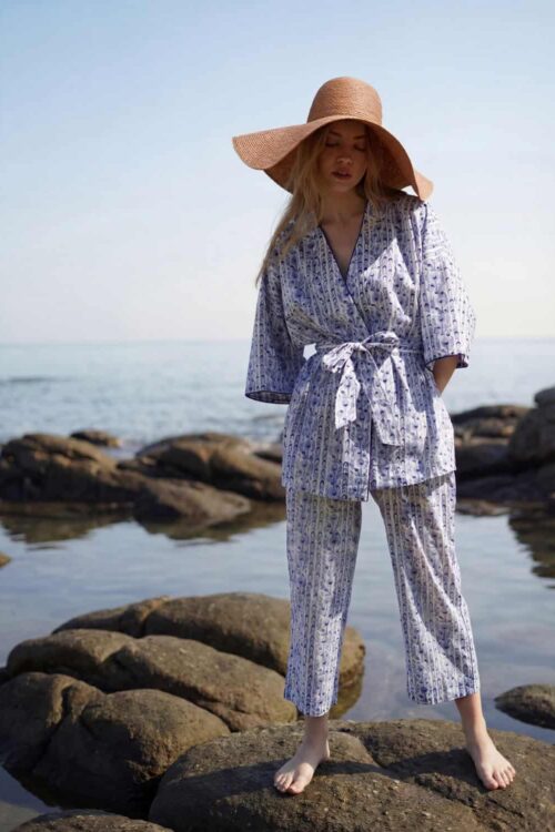 Woman in floral pajamas and wide-brim hat standing on rocks by the sea on a cloudy day.