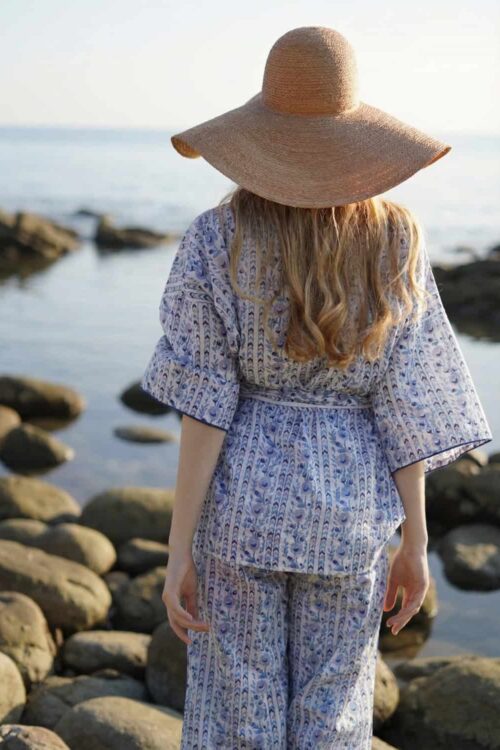 Woman in a wide-brimmed straw hat and patterned outfit standing on rocky shoreline during cloudy wea.