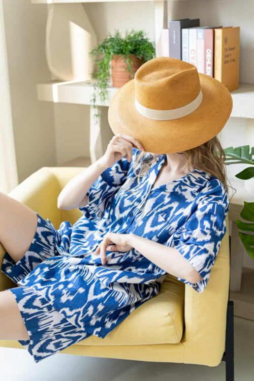 Stylish woman wearing a wide-brimmed hat and blue patterned dress, sitting on a yellow sofa in a coz.