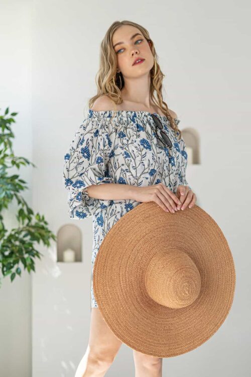 Woman holding a large straw hat, wearing a floral off-shoulder dress, standing indoors with a plant.