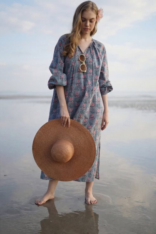 Woman in a floral dress holding a large straw hat at the beach.