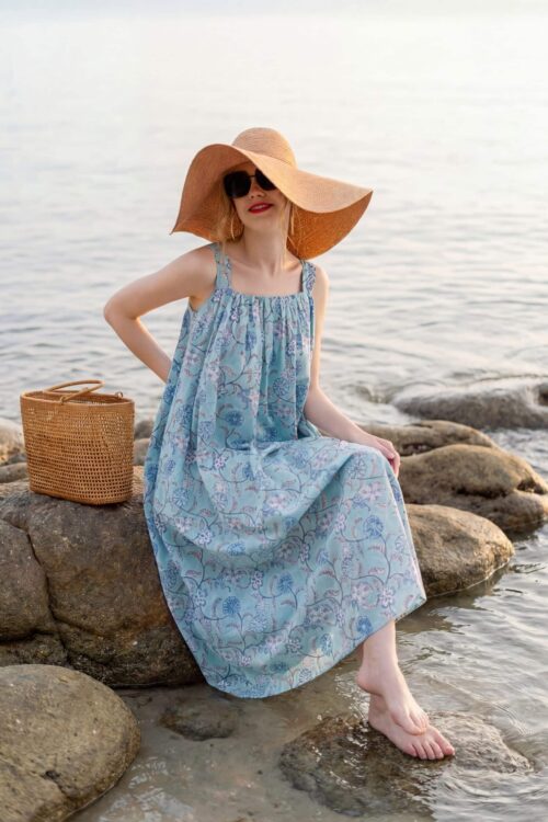 Woman in blue summer dress sitting on rocks by the water with a straw hat.