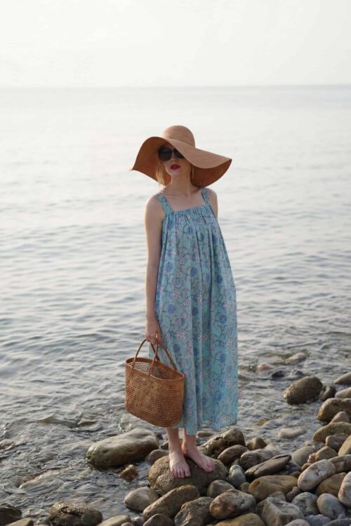 Woman standing on rocky shoreline wearing a blue summer dress, large sun hat, and sunglasses by the.
