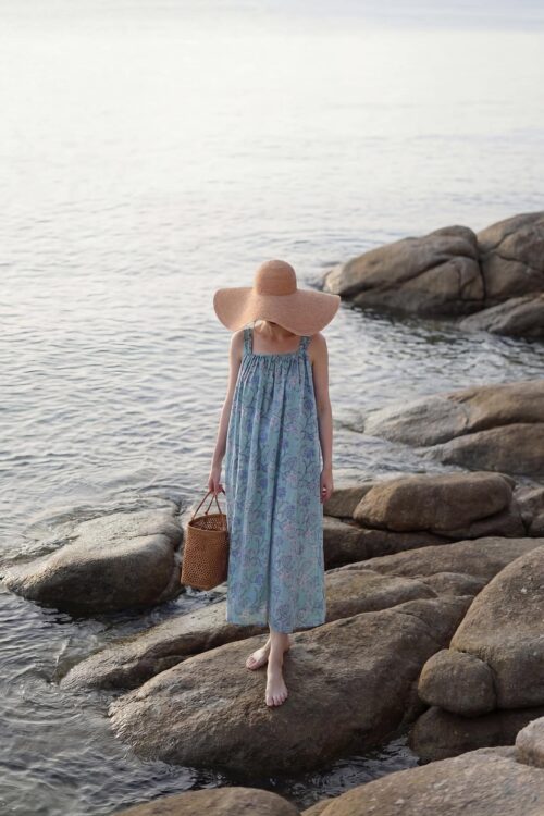 Woman in a blue dress and large sun hat walking on rocks by the sea.