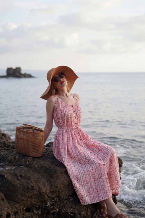 Woman in pink dress with sun hat sitting on rocks by the ocean.