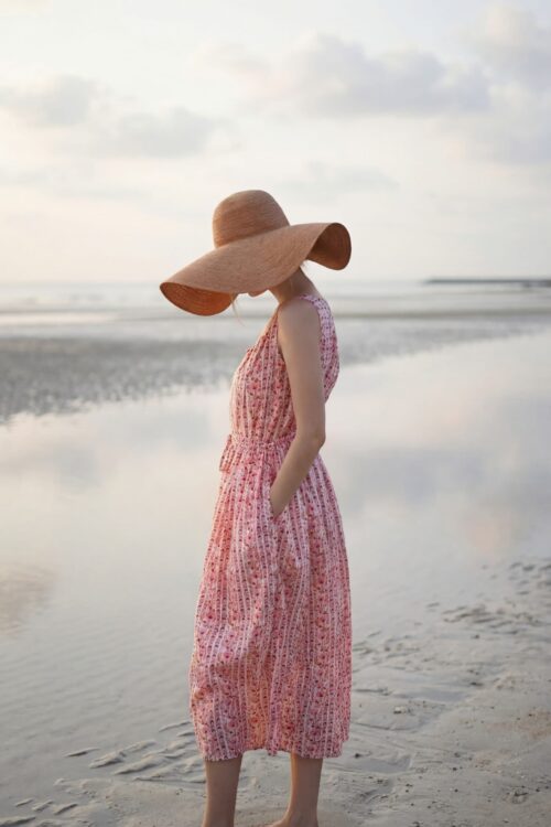 Woman in pink patterned dress and large straw hat standing on the beach.