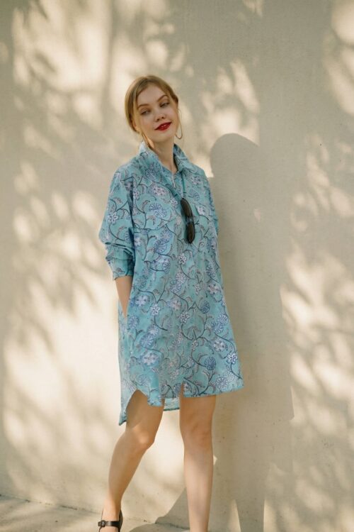 Woman in a blue patterned dress with sunglasses hanging from the collar, standing against a light-co.