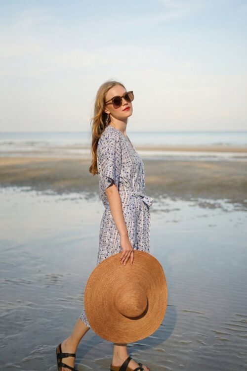Stylish woman in a floral dress holding a straw hat at the beach during April.