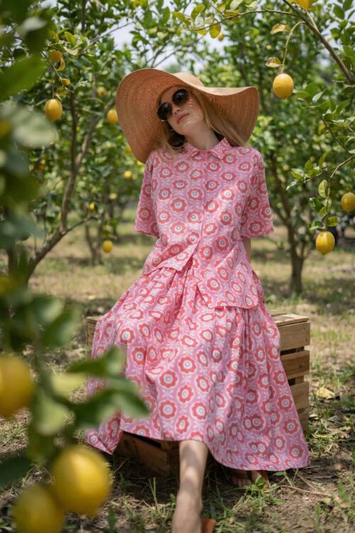 Woman in pink floral dress with large hat among lemon trees.