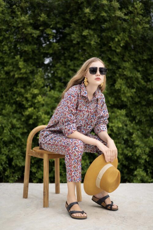 Stylish woman wearing a floral patterned outfit with sunglasses, sitting on a wooden chair outdoors.