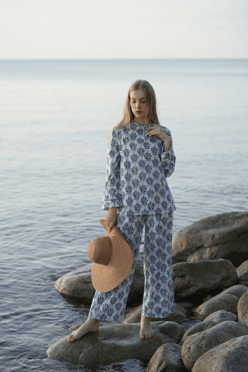 Woman in blue floral bubble pajamas standing on rocks by the sea, holding a straw hat.