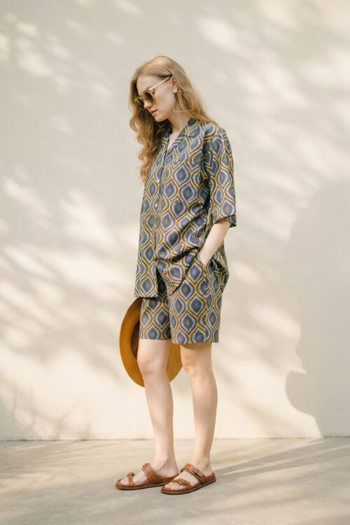 Stylish woman in patterned summer dress with sunglasses and sandals, holding a hat, standing outdoor.