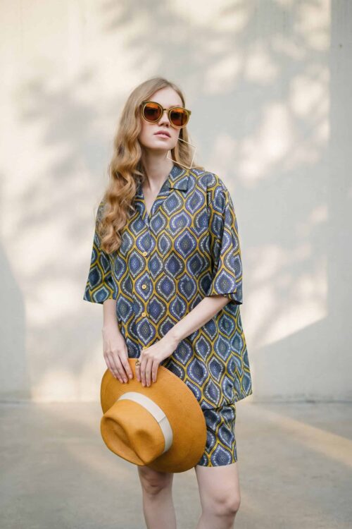 Elegant woman wearing a patterned dress, sunglasses, and holding a hat, showcasing a chic summer loo.