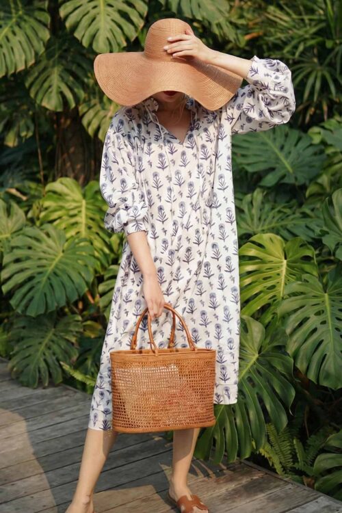 Woman in floral dress with large sun hat and wicker bag in lush tropical setting.