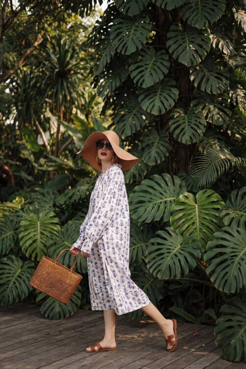 Woman in floral dress with wide-brim hat walking through tropical plants.