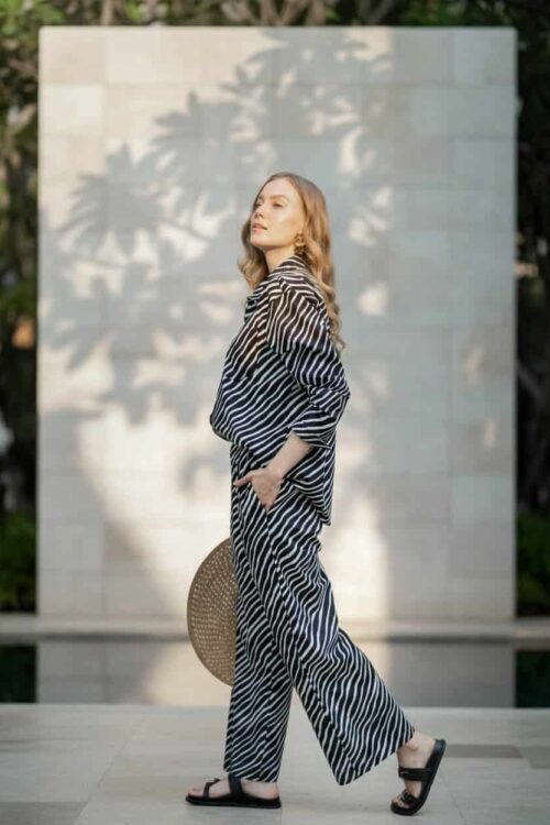 Woman in striped outfit holding a hat, standing outdoors in sunlight.