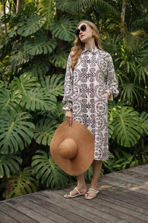 Elegant woman in a patterned dress holding a wide-brimmed straw hat, standing amidst lush green trop.
