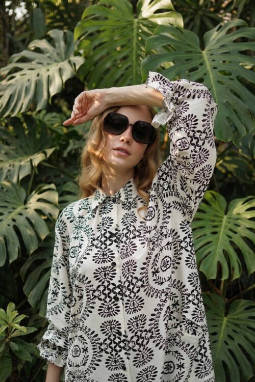 Woman wearing sunglasses and a patterned blouse in a lush garden setting with large Monstera leaves.