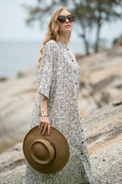 Woman in a patterned summer dress holding a hat by the seaside.