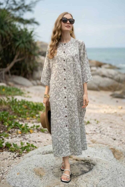 Woman in floral summer dress standing on beach with rocks and greenery.
