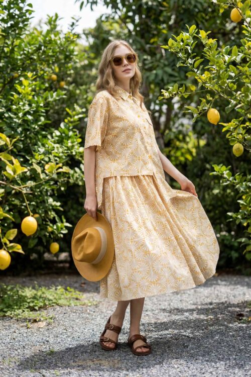 Woman in yellow dress holding hat among lemon trees, sunny day, fashion style.