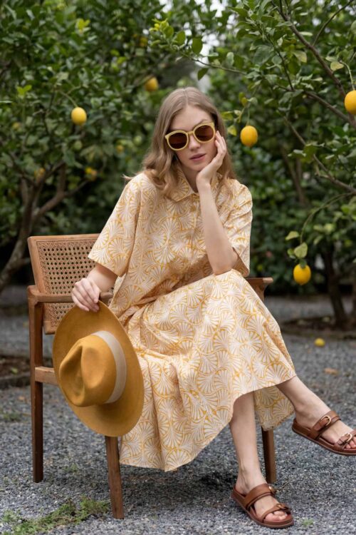 Woman in yellow dress with hat and sunglasses sitting in lemon orchard.