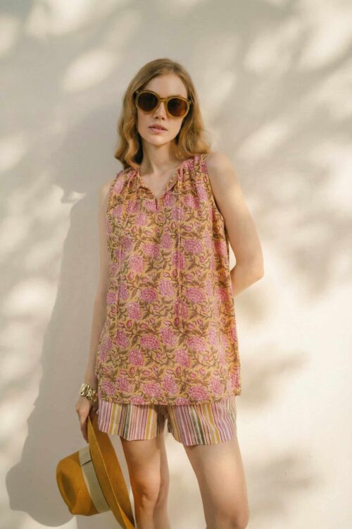 Woman in floral sleeveless top and striped shorts, wearing sunglasses, holding a hat.