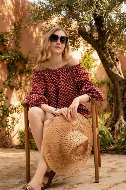 Elegant woman in a red off-shoulder dress with sunglasses sitting outdoors.