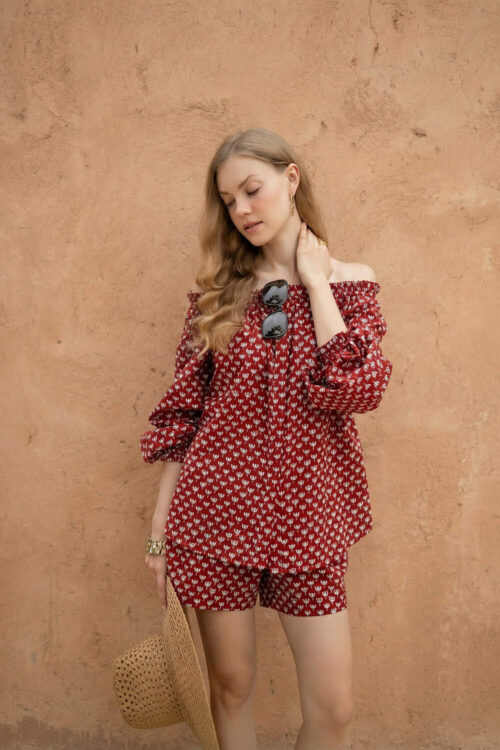 Woman in red carrot-patterned blouse and shorts, holding a straw hat against a beige wall.
